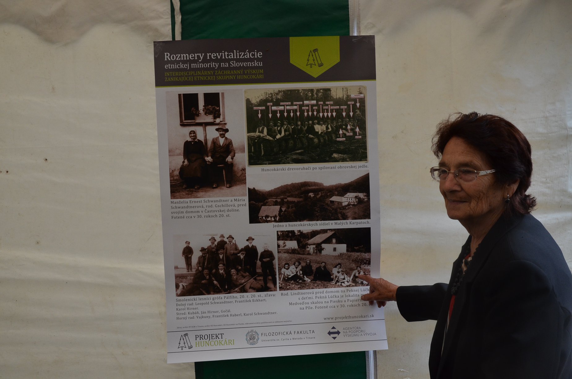 Elderly woman pointing at an exhibition panel of historical Huncokár community photographs