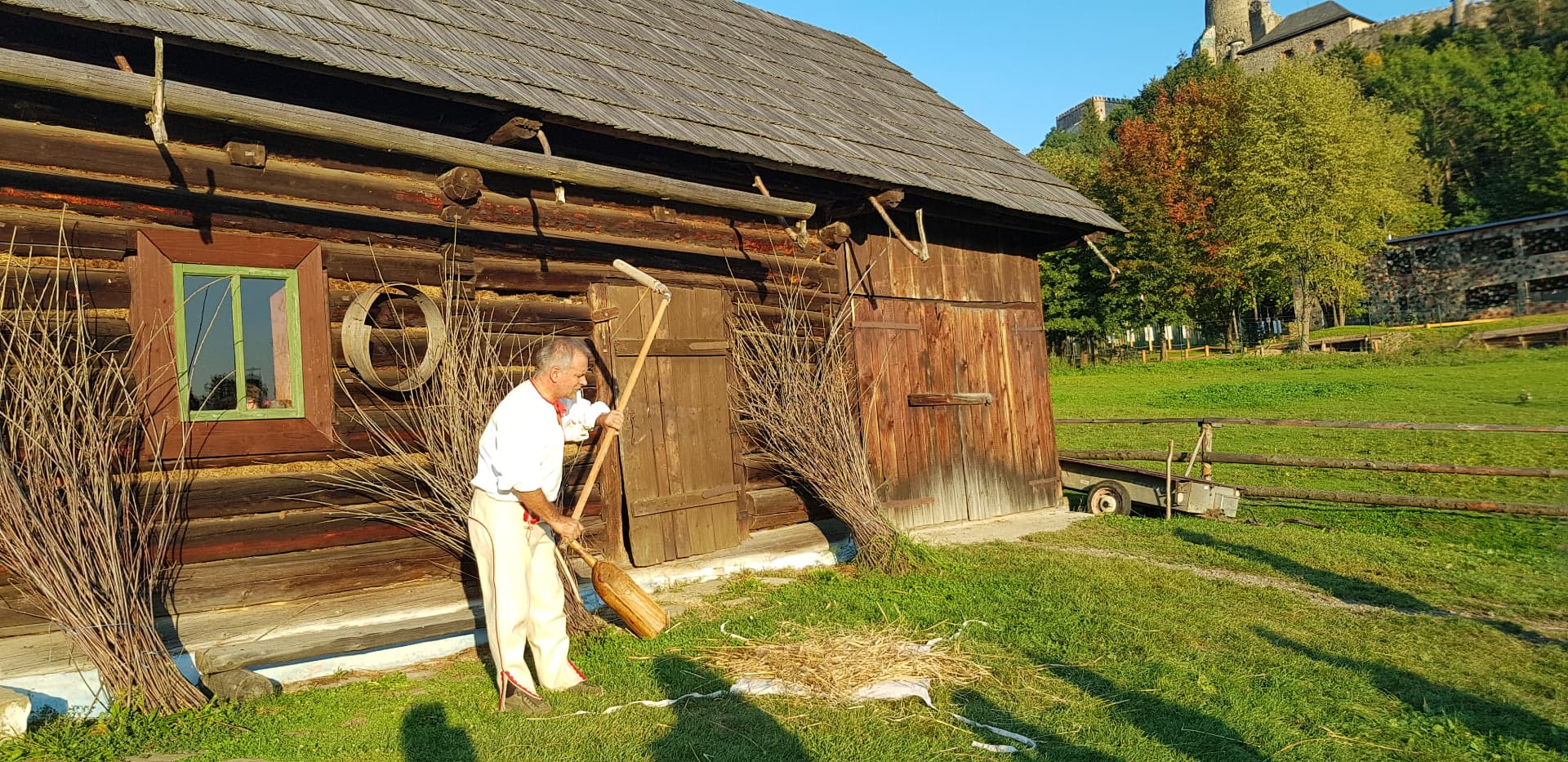Man in traditional dress demonstrating threshing work in front of a wooden rural building